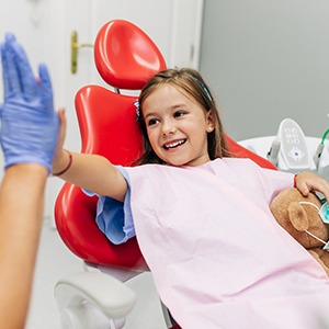 A young girl gives her dentist a high-five while holding a teddy bear and smiling